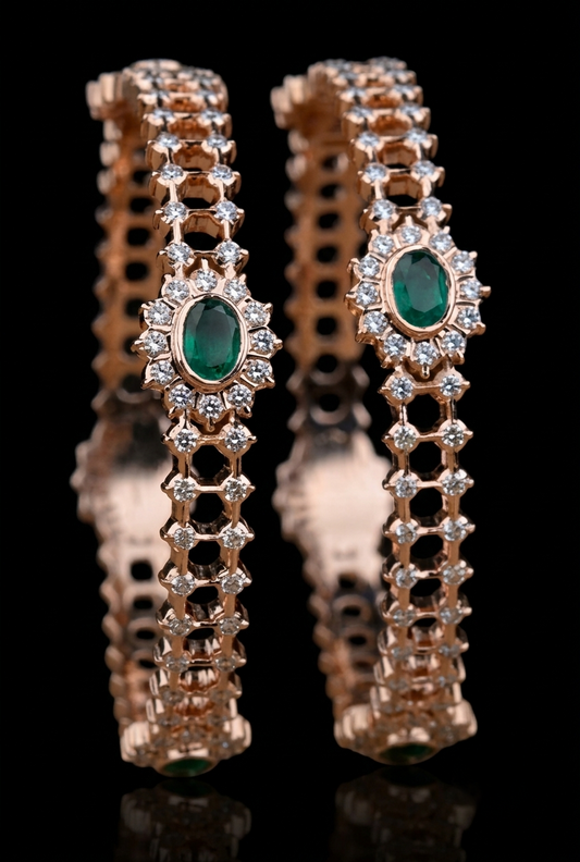 A detailed studio photograph of a pair of gold bangles against a black background. Each bangle features an intricate hexagonal lattice design studded with small round diamonds. The focal point of each is a central oval-cut emerald surrounded by a sparkling diamond halo. A soft reflection of the jewelry is visible beneath them
