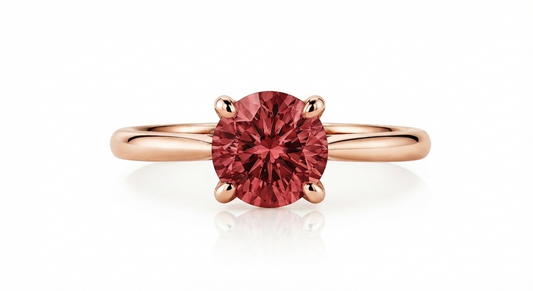 A rose gold solitaire ring featuring a round-cut pinkish-red gemstone in a four-prong setting, shown on a clean white background with a soft reflection.