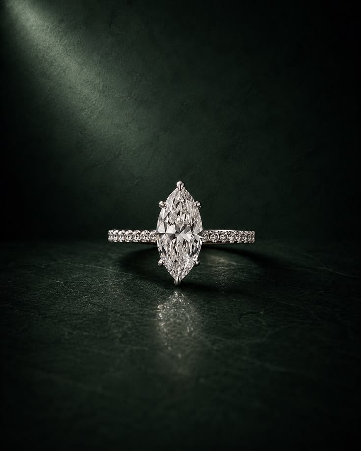 A marquise-cut diamond engagement ring with a pavé band, photographed on a dark textured surface with dramatic spotlighting.