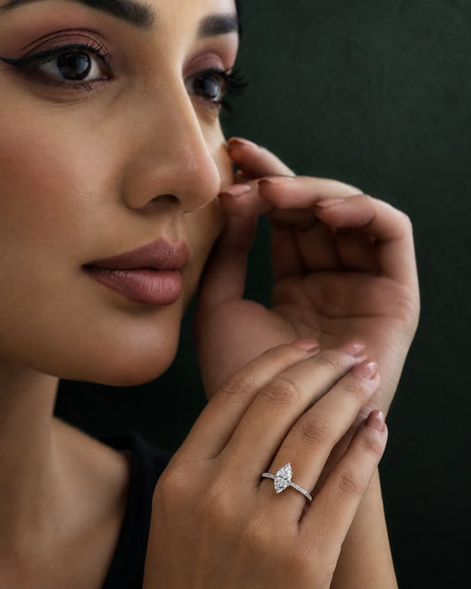 A close-up lifestyle shot of a woman wearing a white gold ring with a marquise-cut solitaire on her finger, set against a dark green background.