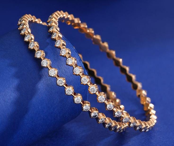A pair of gold tennis-style bangles adorned with clear round stones, Pair of slender rose gold single-line diamond bangles with a subtle scalloped edge pattern, displayed against a royal blue satin background.