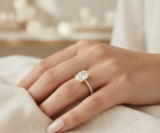 A close-up of a hand wearing a gold ring with a large oval clear gemstone and a pavé band, resting on soft beige fabric.