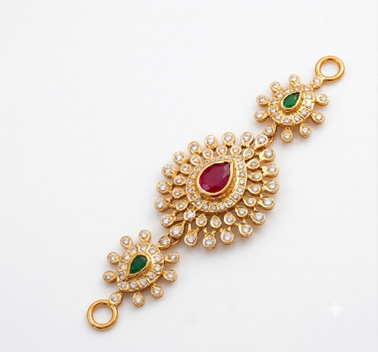 Gold traditional Indian jewelry piece featuring a central teardrop-shaped red ruby stone surrounded by clear pavé stones, with two smaller floral clusters at the ends, each accented with a green emerald-cut stone, all on a plain white background.