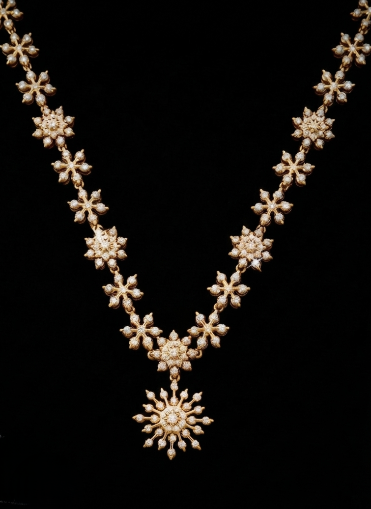 A gold DIAMOND necklace with repeating snowflake-shaped clusters of small clear stones, arranged in a V shape with a larger sunburst pendant at the center, shown against a solid black background.