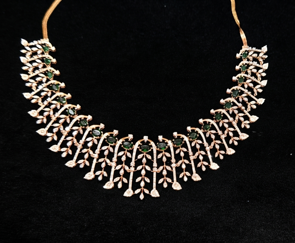 A gold-toned statement necklace featuring intricate leaf and vine motifs embellished with small clear stones and green gemstone centers, displayed against a black background.