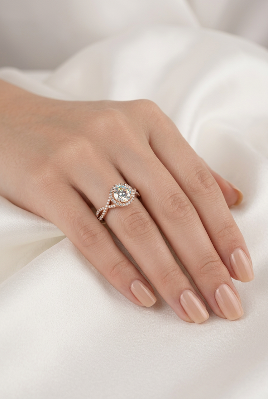 A close-up of a hand with nude manicured nails wearing a delicate rose gold ring with a central clear gemstone and small accent stones, set against a soft white background.